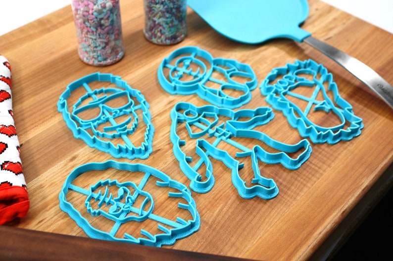 May include: A set of six blue plastic cookie cutters shaped like different cartoon characters. The cutters are on a wooden surface with two jars of sprinkles in the background.