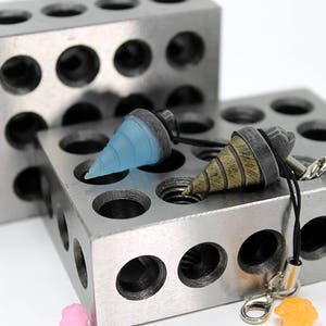 May include: Two drill bit charms on a black cord, one is light blue and the other is brown and gold. The charms are displayed on a silver metal block with holes.