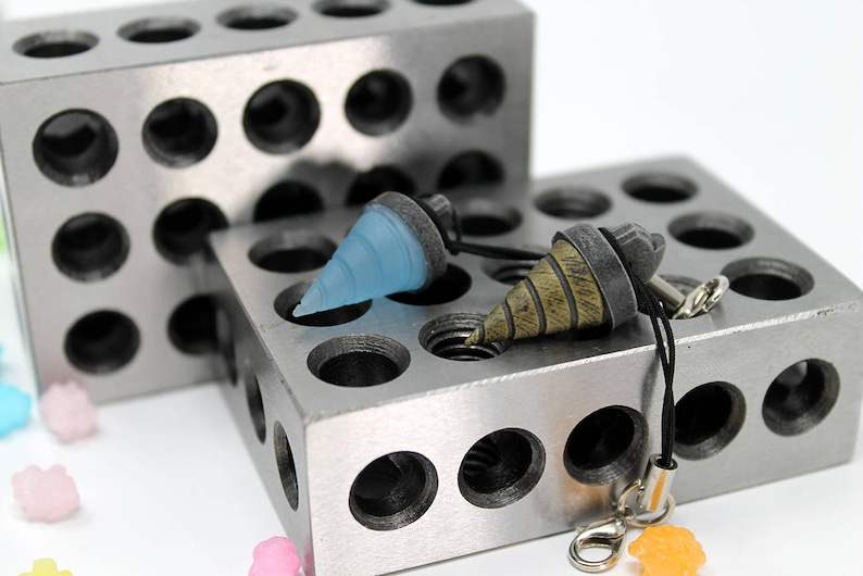 May include: Two drill bit charms on a black cord, one is light blue and the other is brown and gold. The charms are displayed on a silver metal block with holes.