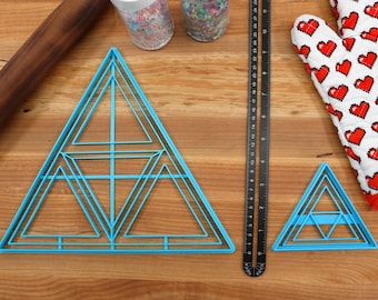 Puede incluir: Dos cortadores de galletas de plástico azul en forma de triángulo sobre una superficie de madera. El cortador más grande tiene varias capas de triángulos, mientras que el más pequeño tiene tres.