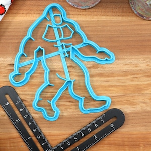 May include: A blue plastic cookie cutter shaped like a Bigfoot walking. The cutter is about 6 inches tall and 4 inches wide. It is sitting on a wooden surface.