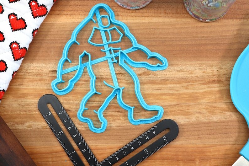 May include: A blue plastic cookie cutter shaped like a Bigfoot walking. The cutter is about 6 inches tall and 4 inches wide. It is sitting on a wooden surface.