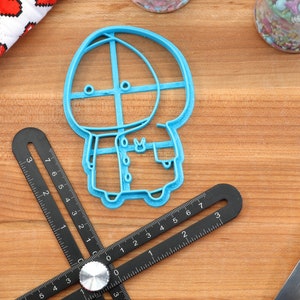 May include: A blue plastic cookie cutter shaped like a cartoon character wearing a hooded coat. The character has a round head with two eyes and a small mouth.