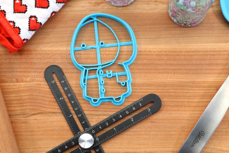 May include: A blue plastic cookie cutter shaped like a cartoon character wearing a hooded coat. The character has a round head with two eyes and a small mouth.