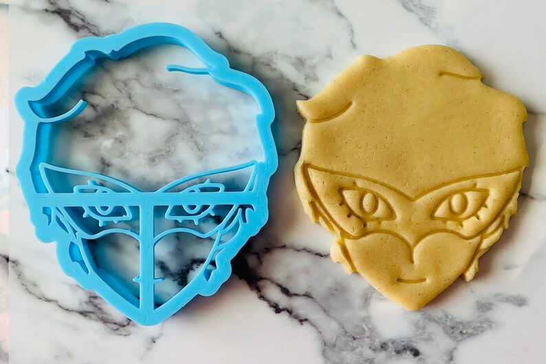 May include: A blue plastic cookie cutter shaped like a cartoon character with a mask and large eyes, next to a light brown cookie cut out in the same shape.
