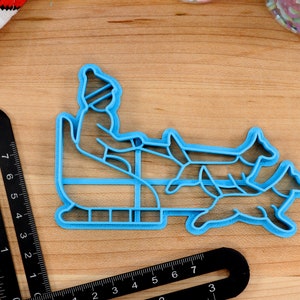 May include: Blue plastic cookie cutter shaped like a person riding a sled pulled by two dogs. The person is wearing a hat and mittens.