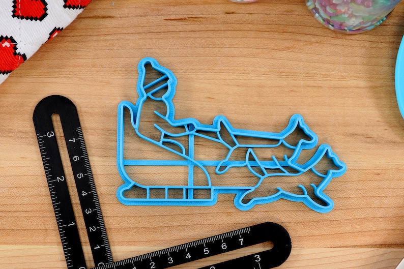 May include: Blue plastic cookie cutter shaped like a person riding a sled pulled by two dogs. The person is wearing a hat and mittens.