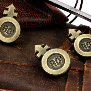 May include: Three antique gold-toned metal tokens with a symbol and the letters "AD" and the number "2" on each. The tokens are on a brown leather surface.