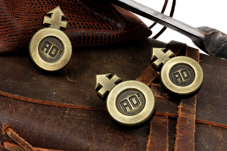 May include: Three antique gold-toned metal tokens with a symbol and the letters "AD" and the number "2" on each. The tokens are on a brown leather surface.