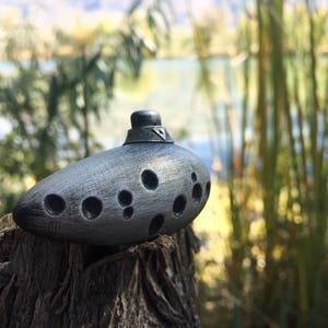 Puede incluir: Una ocarina plateada con un dise&ntilde;o triangular en la parte superior y varios agujeros en el lateral, sentada sobre un toc&oacute;n de &aacute;rbol desgastado.