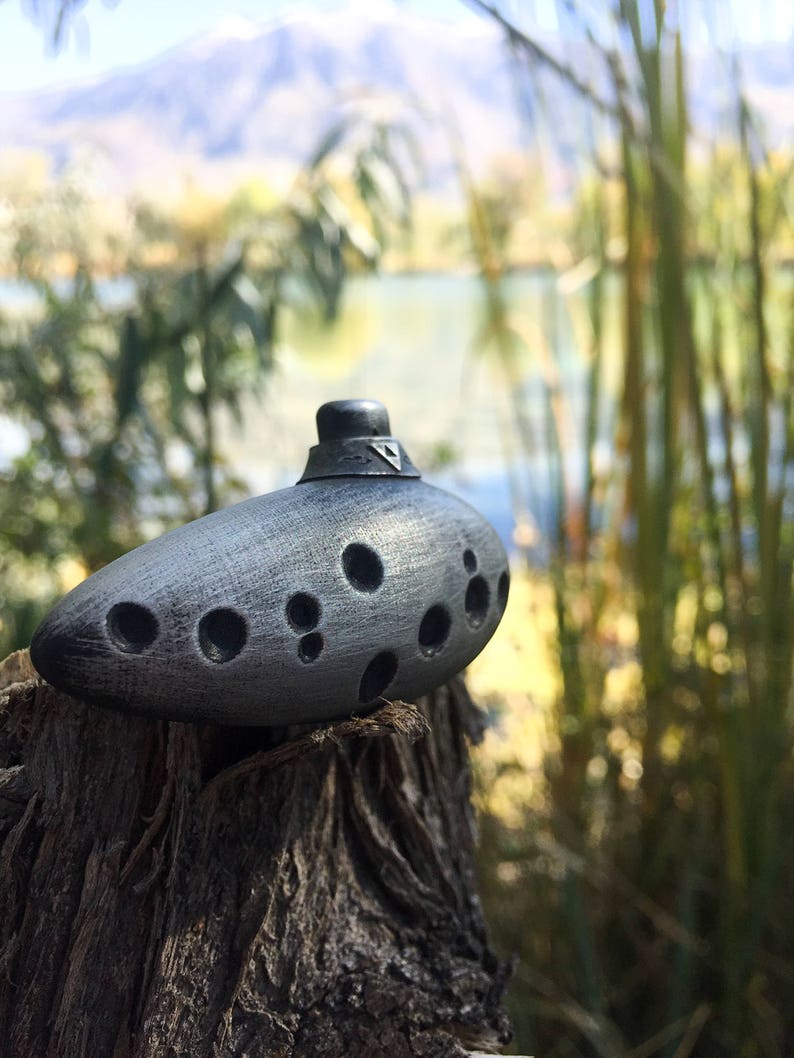 Puede incluir: Una ocarina plateada con un dise&ntilde;o triangular en la parte superior y varios agujeros en el lateral, sentada sobre un toc&oacute;n de &aacute;rbol desgastado.