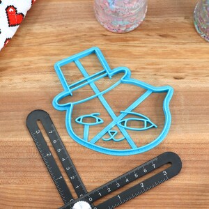 May include: A blue plastic cookie cutter shaped like a cat wearing a top hat. The cookie cutter is on a wooden surface.