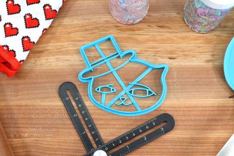 May include: A blue plastic cookie cutter shaped like a cat wearing a top hat. The cookie cutter is on a wooden surface.