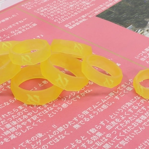 May include: A group of eight yellow rubber rings with a flame design. The rings are scattered on a pink surface.