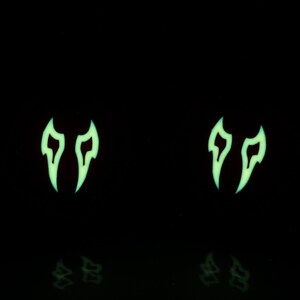 May include: Two glow-in-the-dark green flame-shaped earrings. The earrings are on a black background.