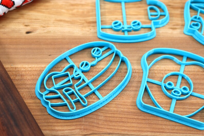 May include: A set of blue plastic cookie cutters shaped like a coffee cup, a donut, a hamburger, and a loaf of bread. The cutters are on a wooden surface.