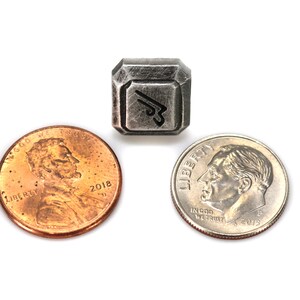 May include: A silver-colored metal charm with a stylized design, similar in size to a US penny.