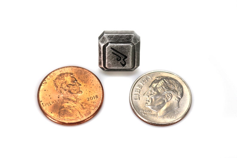 May include: A silver-colored metal charm with a stylized design, similar in size to a US penny.