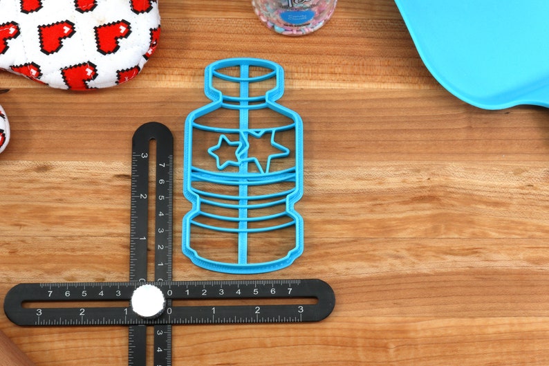 Puede incluir: Un cortador de galletas de pl&aacute;stico azul con forma de botella de agua con dos estrellas en la parte delantera. El cortador mide 17,8 cm de alto y est&aacute; sobre una superficie de madera.
