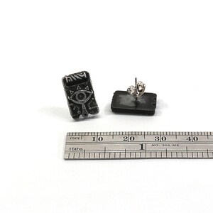 May include: A pair of silver stud earrings featuring a black and white image of a Triforce symbol with an eye in the center. The earrings are approximately 1 cm in diameter.