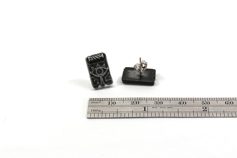 May include: A pair of silver stud earrings featuring a black and white image of a Triforce symbol with an eye in the center. The earrings are approximately 1 cm in diameter.