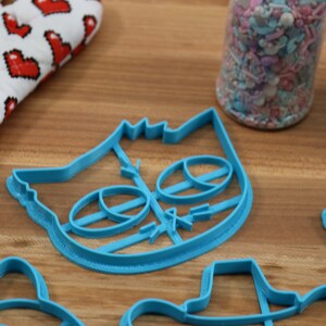 May include: A set of blue plastic cookie cutters, including a cat face, a unicorn, a hat, and a flower. The cutters are on a wooden surface.