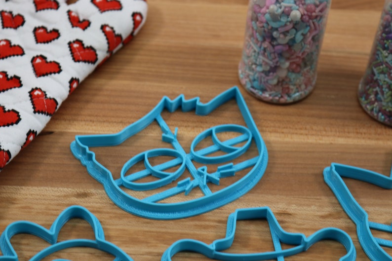May include: A set of blue plastic cookie cutters, including a cat face, a unicorn, a hat, and a flower. The cutters are on a wooden surface.