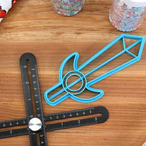 May include: A blue plastic sword cookie cutter on a wooden surface. The cutter is shaped like a sword with a rounded handle and a pointed tip.