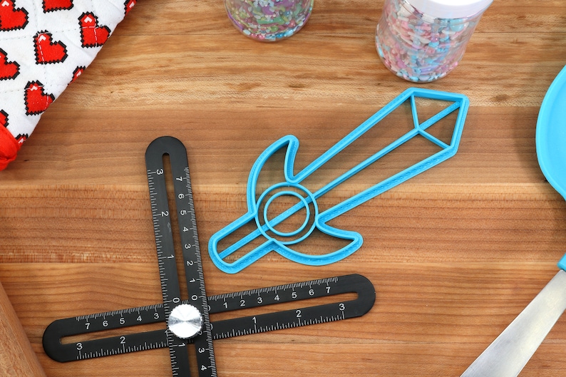 May include: A blue plastic sword cookie cutter on a wooden surface. The cutter is shaped like a sword with a rounded handle and a pointed tip.