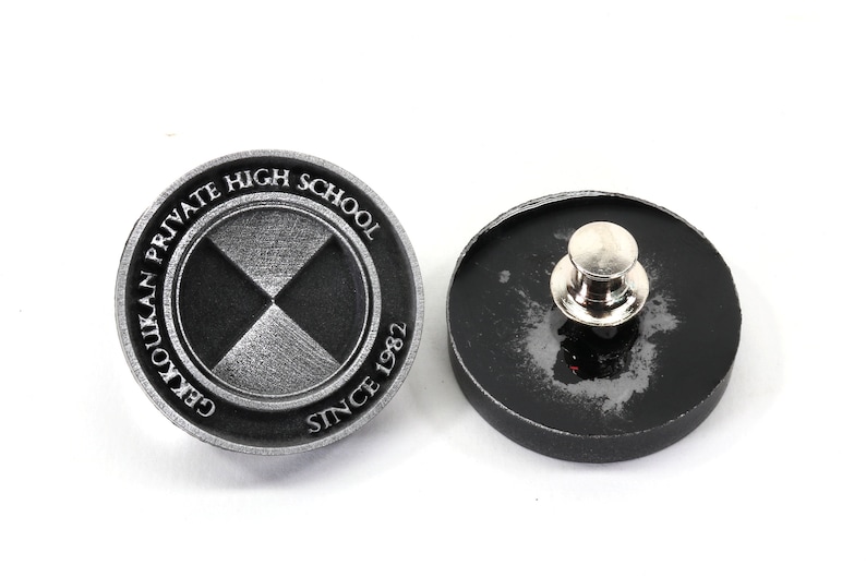 Pu&ograve; includere: Uno spillo argentato con un designo bianco e nero. Lo spillo presenta il testo "Gekkoukan Private High School Since 1982". Il retro dello spillo &egrave; nero.