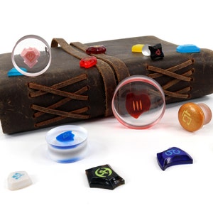 May include: A collection of colorful glass and plastic dice with various symbols and designs. The dice are scattered on a white surface next to a brown leather-bound journal with a leather strap.