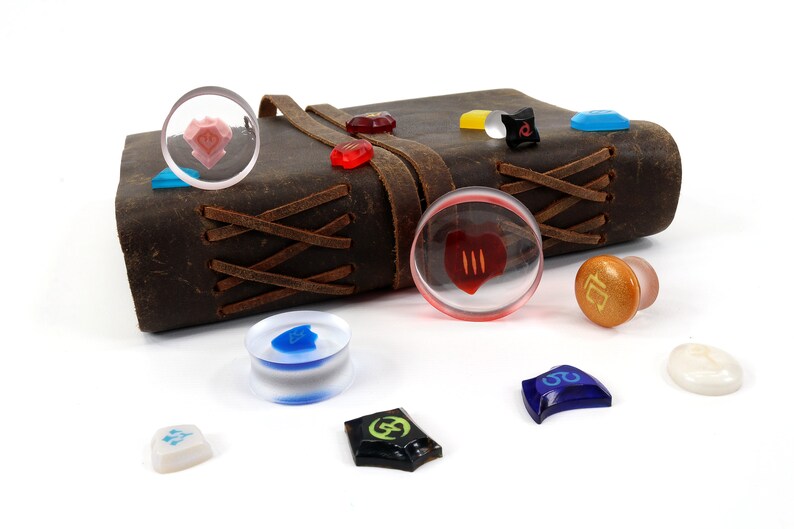 May include: A collection of colorful glass and plastic dice with various symbols and designs. The dice are scattered on a white surface next to a brown leather-bound journal with a leather strap.