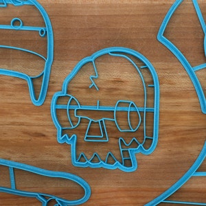 May include: A set of five blue plastic cookie cutters shaped like a skateboard, a skull, a hat, a shirt, and a box. The cutters are on a wooden surface.