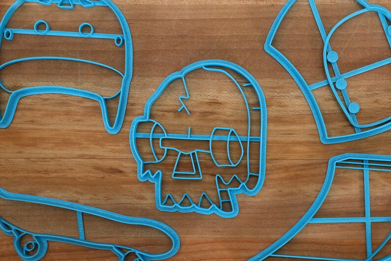 May include: A set of five blue plastic cookie cutters shaped like a skateboard, a skull, a hat, a shirt, and a box. The cutters are on a wooden surface.