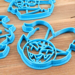 May include: A set of four blue plastic cookie cutters shaped like a pirate ship, a treasure chest, a skull and crossbones, and a mermaid. The cutters are on a wooden surface.