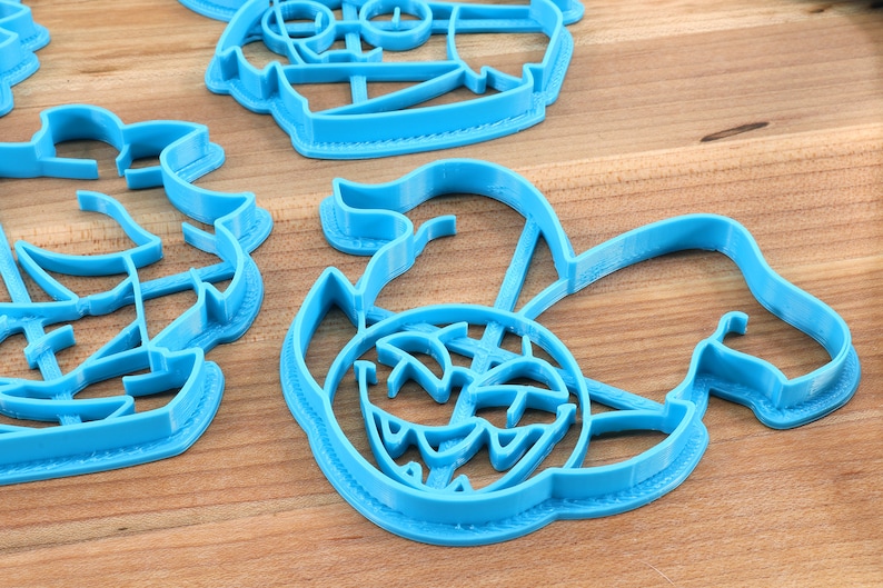 May include: A set of four blue plastic cookie cutters shaped like a pirate ship, a treasure chest, a skull and crossbones, and a mermaid. The cutters are on a wooden surface.