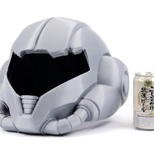 May include: A gray 3D printed helmet with a large visor and intricate details. The helmet is sitting on a white surface next to a can of beer.