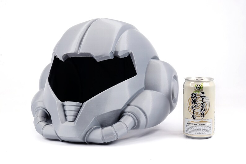 May include: A gray 3D printed helmet with a large visor and intricate details. The helmet is sitting on a white surface next to a can of beer.