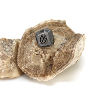 May include: A small, dark grey, geometrically shaped stone with a symbol etched into it sits on top of a larger, light brown, layered rock.