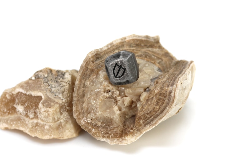 May include: A small, dark grey, geometrically shaped stone with a symbol etched into it sits on top of a larger, light brown, layered rock.