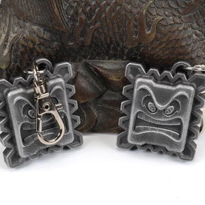 May include: Two silver-colored keychains with a detailed, angry face design. The keychains are attached to a silver keyring.