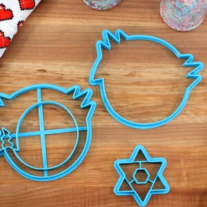 May include: Three blue plastic cookie cutters on a wooden surface. The cutters are shaped like a star, a circle with a cross inside, and a rounded shape with a jagged edge.