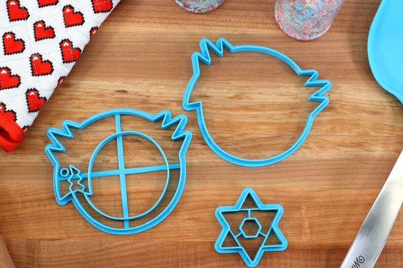 May include: Three blue plastic cookie cutters on a wooden surface. The cutters are shaped like a star, a circle with a cross inside, and a rounded shape with a jagged edge.