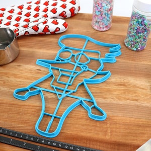 May include: A blue plastic cookie cutter shaped like a cartoon character wearing a hat. The cutter is on a wooden surface with a rolling pin, measuring cup, and two jars of sprinkles.