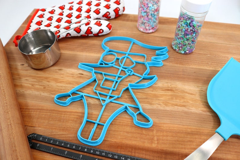 May include: A blue plastic cookie cutter shaped like a cartoon character wearing a hat. The cutter is on a wooden surface with a rolling pin, measuring cup, and two jars of sprinkles.