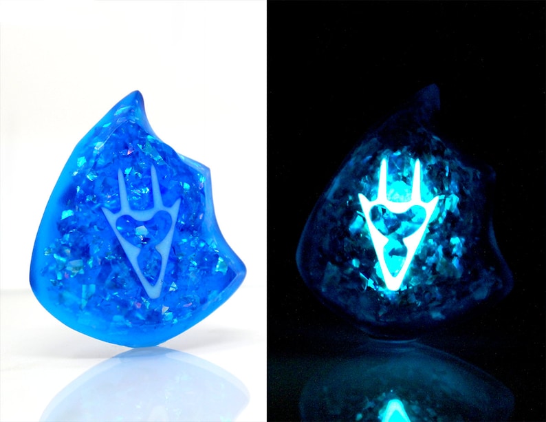 May include: A blue resin pendant with a white, glowing symbol inside. The pendant is filled with iridescent glitter and glows in the dark.