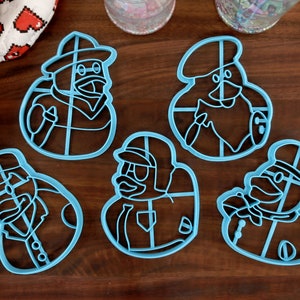 Puede incluir: Cinco cortadores de galletas de plástico azul con forma de pingüinos que llevan sombreros y bufandas. Los cortadores están sobre una superficie de madera.