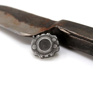 May include: A silver-colored metal pin with a geometric design. The pin has a square in the center and is surrounded by a circle with a series of smaller circles around it. The pin is lying on a white surface.