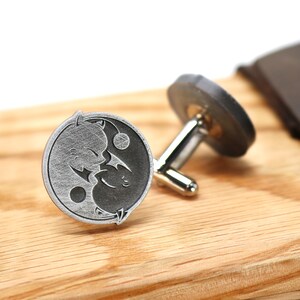 May include: A pair of silver cufflinks with a yin yang design featuring two stylized cats. The cufflinks are on a wooden surface.