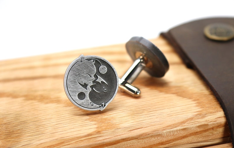 May include: A pair of silver cufflinks with a yin yang design featuring two stylized cats. The cufflinks are on a wooden surface.